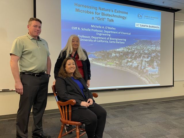 Professor Michelle A. O’Malley sitting in a chair with two other people at her sides in front of her presentation board.